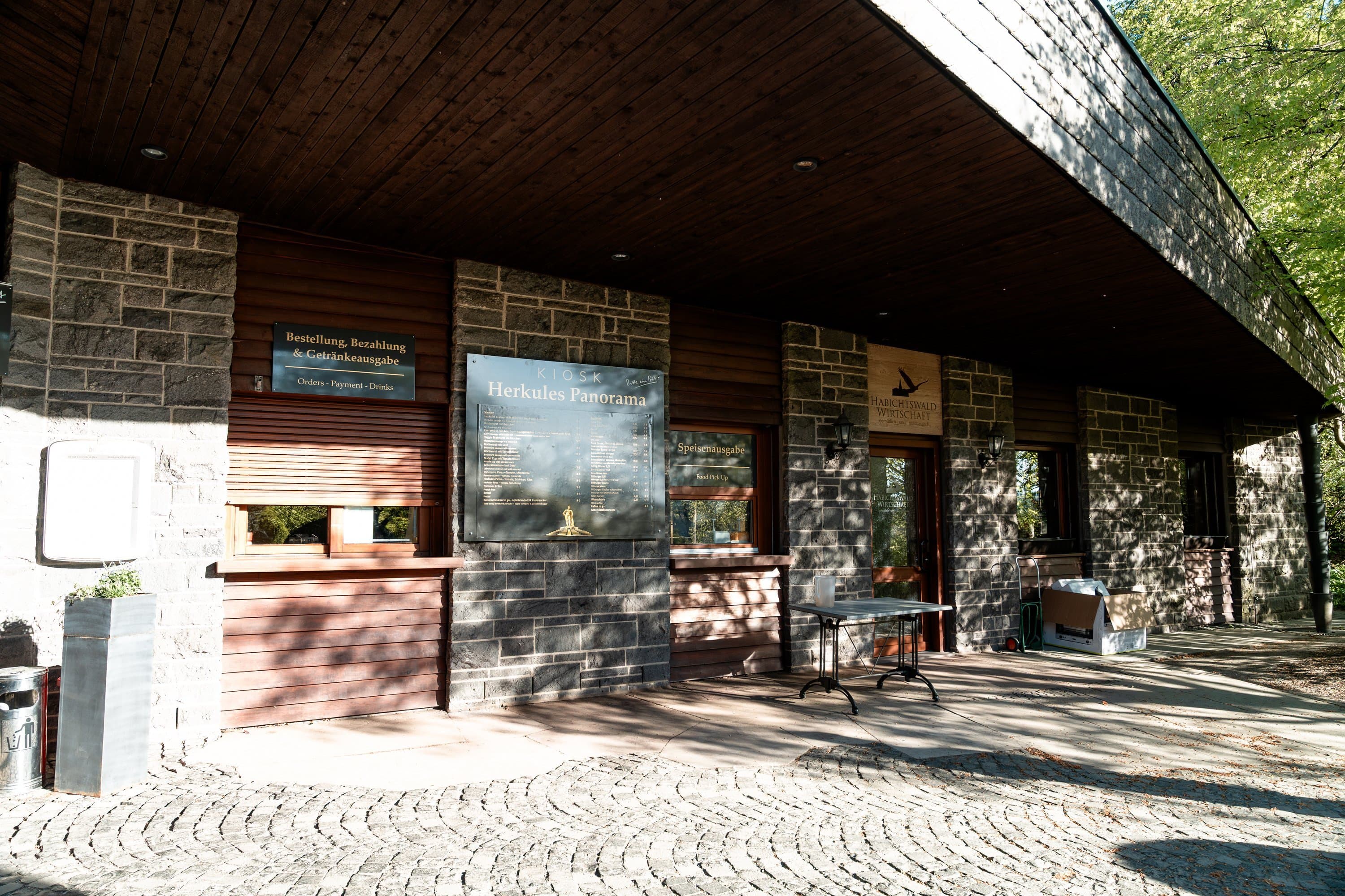 Außenansicht des Kiosks am Herkules Panorama mit Tischen und Sonnenschirmen.
